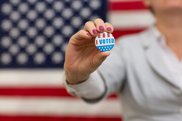 Voter woman holds in hand an USA badge at vote center closeup. Woman stands in front of U.S. flag on presidential election day. Motivation of a political campaign. I voted badge in womand hand closeup