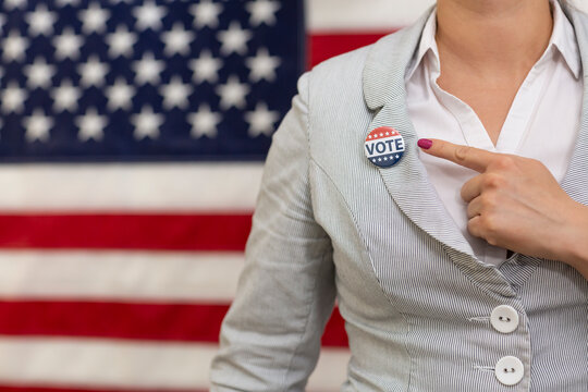 Young voter woman  waist up pointing with finger to usa badge at vote center. Woman stands U.S. flag in background at electoral college presidential election day. Motivation of a political campaign. 