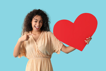 Beautiful young African-American woman with red paper heart pointing at something on blue background