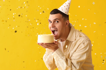 Young man in party hat with sweet cake and confetti celebrating Birthday on yellow background