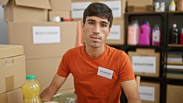 Devoted young hispanic man actively volunteering at a charity center, sitting at the table with a serious face, working diligently in a room full of donations