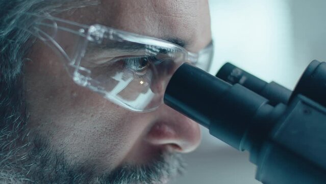 Mature male scienting with gray hair and beard wearing protective glasses looking through microscope in laboratory. Close-up view, tilt up shot