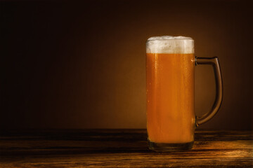 glass of cool unfiltered wheat beer with head of foam stands on a wooden table on a soft background with a gradient light spot. side view with copy space. selective focus