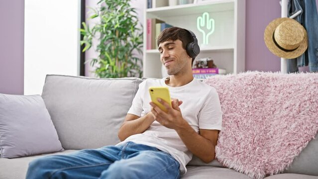 Confident young hispanic man lounges on the living room sofa, grooving to the sound of his favorite songs. his mobile turned dance floor, he lets music fill his home with positive vibes.