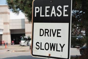 please drive slowly caption writing text rectangle metal sign with parking lot behind, white and...