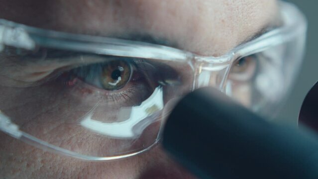 Eyes of mature male chemist in safety glasses looking through microscope during laboratory research. Extreme close-up view
