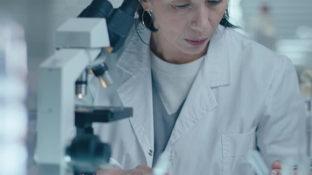 Mid-aged female scientist examining sample under microscope and writing down results during laboratory test. Tilt-down shot