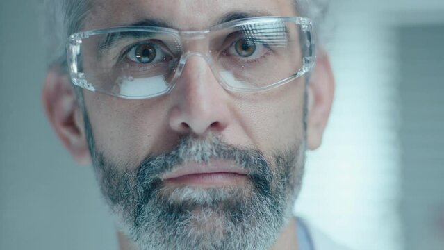Mature male scientist with gray hair and beard wearing safety glasses posing on camera in laboratory. Close up shot, video portrait