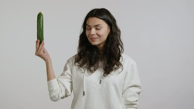 A young black-haired girl is promoting a beautiful green cucumber. She is in a white hoodie against a white background. The girl is happy and joyful. High quality 4k footage