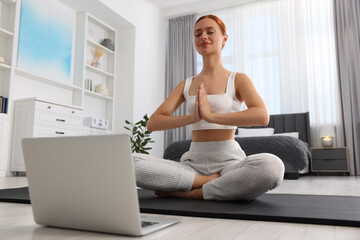 Beautiful young woman practicing Padmasana with laptop on yoga mat at home, low angle view. Lotus pose