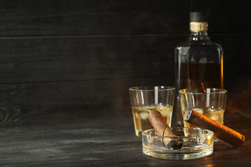 Cigars, ashtray and whiskey with ice cubes on black wooden table. Space for text