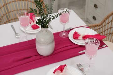 Table setting. Glasses of tasty beverage, plates, pink napkins and vase with green branches in dining room