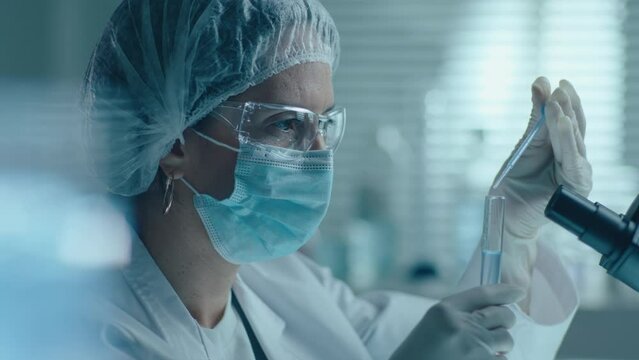 Female scientist in protective mask, gloves and eyewear dropping liquid substance with pipette into test tube, shaking and examining reaction, developing vaccine in lab. Tilt-up shot