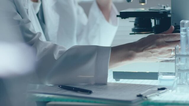 Hands of female scientist adjusting microscope, conducting research and taking notes on clipboard in medical laboratory. Close-up shot