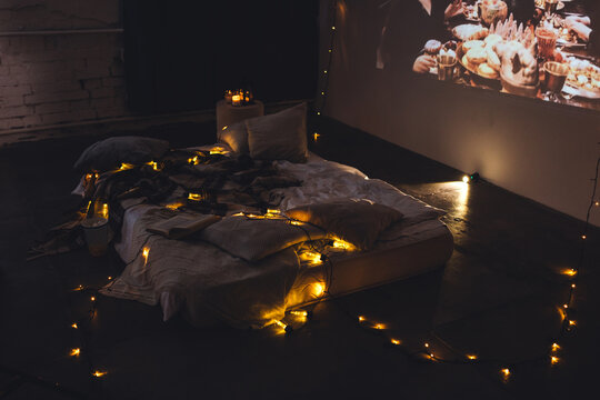 Romantic surprise for girlfriend or boyfriend. Lazy weekend in bed concept. Bedroom prepared for watching old movies with popcorn, decorated with lights and candles. Cozy home atmosphere
