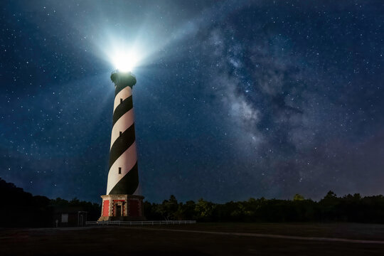 Historic Cape Hatteras Lighthouse shines brightly in a night sky full of stars and the galactic center of the Milky Way Galaxy at Cape Hatteras National Seashore on the Outer Banks of North Carolina.