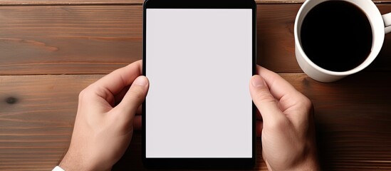 Man's hands holding a black blank screen smartphone, sitting in front of a computer tablet with a white blank screen and stylus pen, on a wooden working desk. - Powered by Adobe