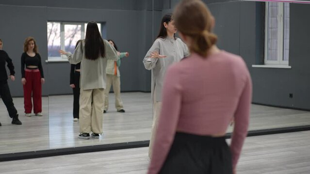 A female instructor conducts group dance exercises in the choreographic hall. Five students carefully watch the teacher's master class