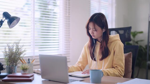 Asian female student engaged in online learning using a laptop and headphones, diligently recording study materials in a somewhat bored manner at her home desk