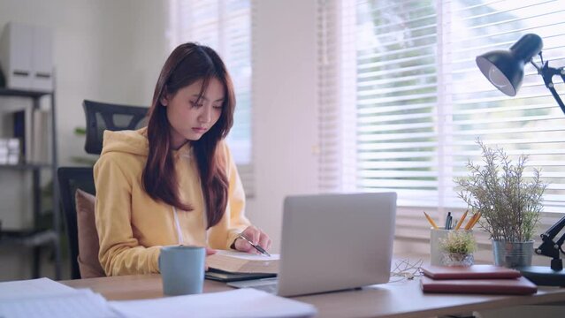 Asian female student engages in focused online learning using her laptop and headphones. She diligently records study materials in a notebook at her home workspace.