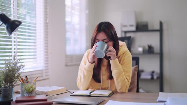 Asian college student reading study materials at her home desk, enjoying a relaxing drink to unwind from her studies for depicting a balanced approach to learning and relaxation
