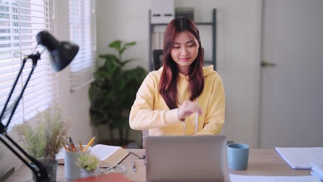 Asian female student engaged in online learning on her laptop at the home desk, battling drowsiness and eventually dozing off