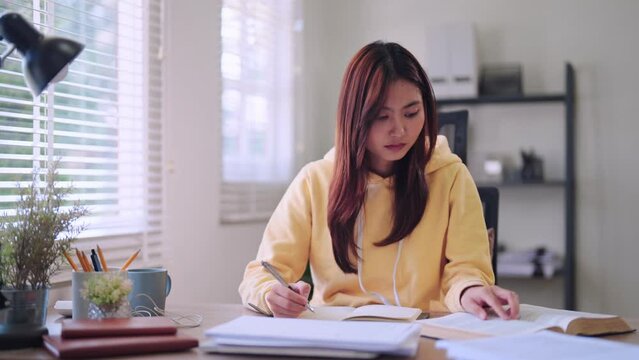 Asian female student immersed in homework and researching study materials through books at her dedicated home desk. Demonstrates focused intent on learning