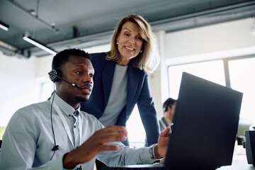 Black businessman with headset working on laptop while female CEO is supervising him in office. - obrazy, fototapety, plakaty