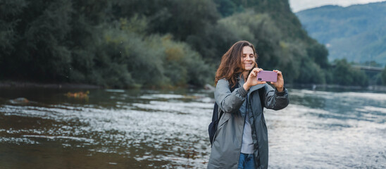 A young woman traveler in a raincoat with a smartphone in her hands on the bank of a mountain rive