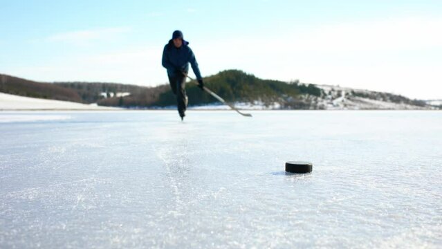 An elderly man practices stricking the puck with hockey stick on a frozen lake in winter.