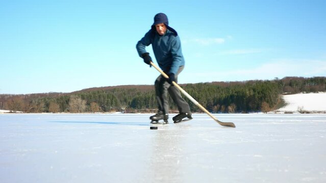 An elderly man practices stricking the puck with hockey stick on a frozen lake in winter.