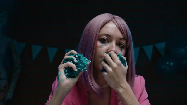 Pink hair birthday girl sitting at the table, taking piece of birthday cake and eating it with her hands