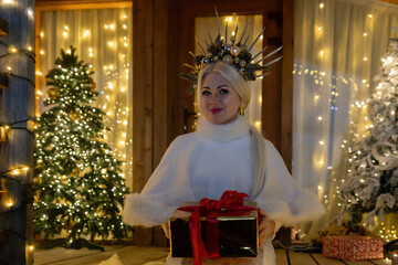 Happy blonde as Snow Maiden in white costume. Wooden house exterior Festive setting. Character with crown creating festive scene.