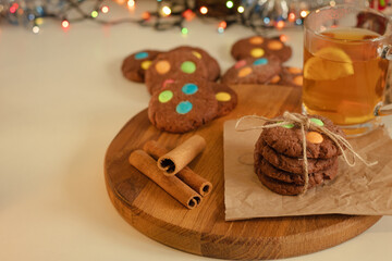 Brown chocolate cookies with small dragees and tea on a wooden board and white table. Blurred bokeh from tinsel and garland. The atmosphere of a home holiday. New Year and Christmas background.
