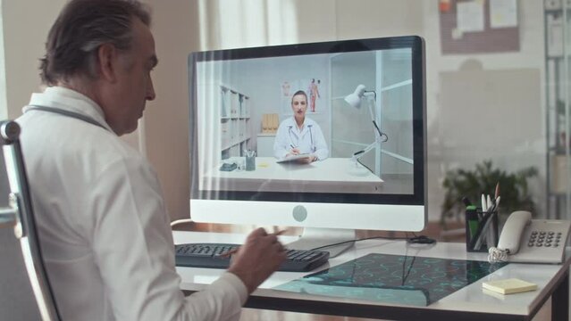 Mature male radiologist in white coat discussing patients X-ray scan with female therapist during video call using computer