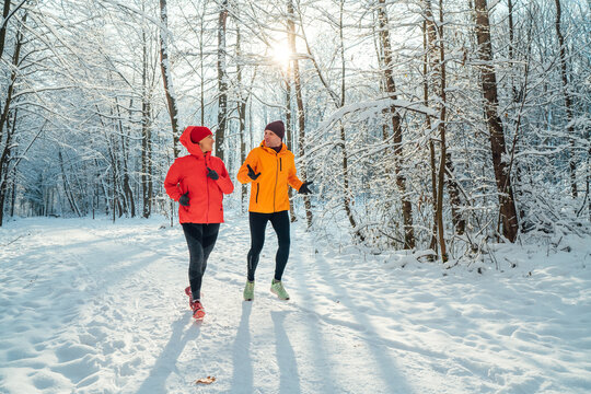 Middle-aged smiling trail couple runners man and woman dressed bright windproof jackets running speaking picturesque snowy forest during sunny frosty day. Sporty active people, winter training image.