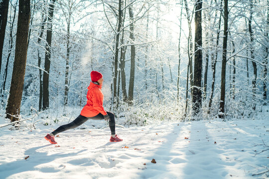 Trail runner woman dressed bright red windproof jacket making stretching exercises in picturesque snowy forest during sunny frosty day. Sporty active people and winter training concept image.