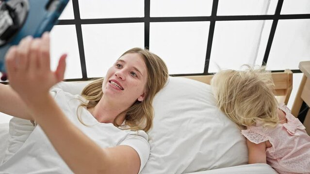 Smiling caucasian mother and sweet little daughter lying together on bed, taking a warm morning selfie using touchpad in cozy bedroom