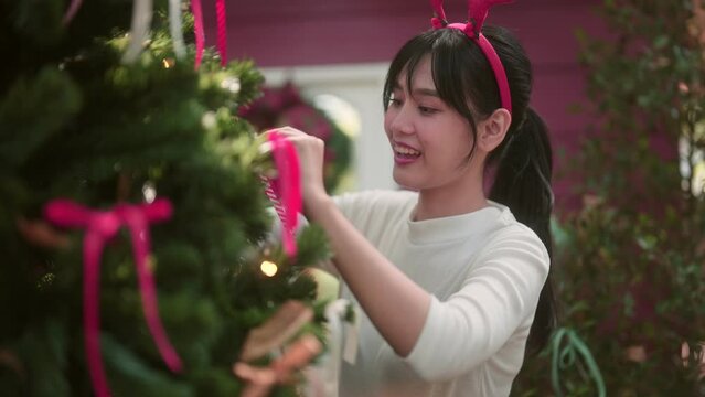 An Asian woman is decorating her Christmas tree, getting ready for the festive season in the cozy, themed ambiance of her home. Creating a warm holiday atmosphere is her joyful preparation