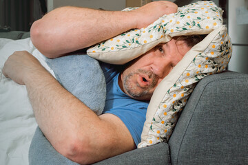 A man covers himself with pillows trying to sleep. Loud sound that disturbs sleep. No peace of mind