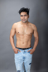 Portrait of a shirtless young man. Healthy muscular man with black hair, posing with bare upper body, hands in pocket on grey background.