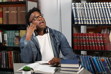 African-American man in eyeglasses sits in office, looking up, talking on cell phone. Against background bookshelves, young black guy is sitting at table with books, opening mouth, looking up