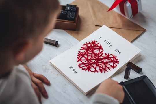 Concept of children craft, handmade gift for Mother's Day. Little boy holding craft card with embroidering in the shape of heart and printed message I love you Mom. Kid's leisure activity, handcraft