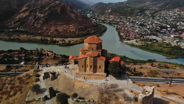 Aerial scenery of amazing city Mtskheta surrounded by rivers and mountains and having old church on the shore. Drone footage of ancient church and tourists walking around it. Cultural heritage theme