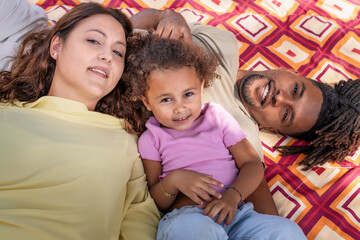 A multiracial family with a young child enjoys a relaxing moment together while laying on a blanket outdoors, showcasing a scene of happiness and familial bond.