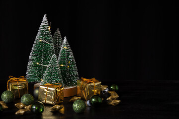 Close-up of green and gold Christmas decoration with little trees with lights on dark table, black background, horizontal, with copy space
