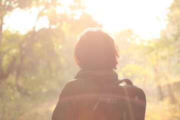 An Indian boy with long hairs wearing backpack and power glasses in front of bright sunlight and sun light flare in the forest, looking towards sun