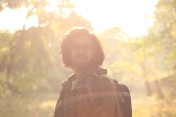 An Indian boy with long hairs wearing backpack and power glasses in front of bright sunlight and...