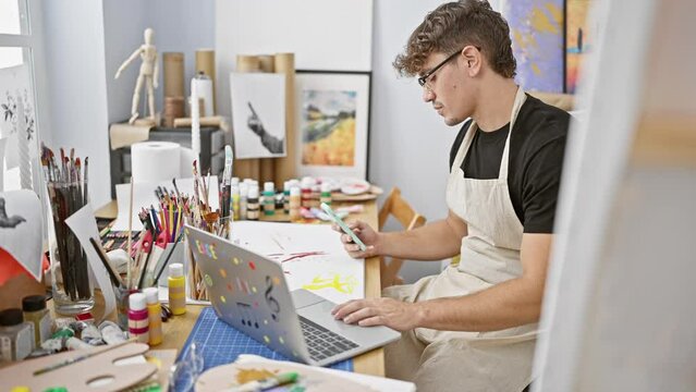 Captivating portrait, serious young hispanic man, an attractive artist, engrossed in learning art online at his indoor studio, using laptop and smartphone amidst paintbrushes and canvas
