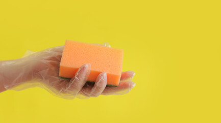 Orange dishwashing sponge in a woman's hand on a yellow background. A hand in a transparent disposable glove holds a sponge for wet cleaning. Professional cleaning. Copy space